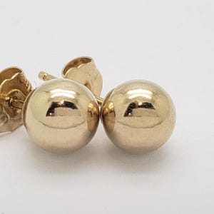 Pu&ograve; includere: Un paio di orecchini a lobo a sfera dorati. Gli orecchini hanno una superficie lucida e riflettente e sono montati su perni d'oro con chiusure a farfalla. Gli orecchini sono piccoli e rotondi.