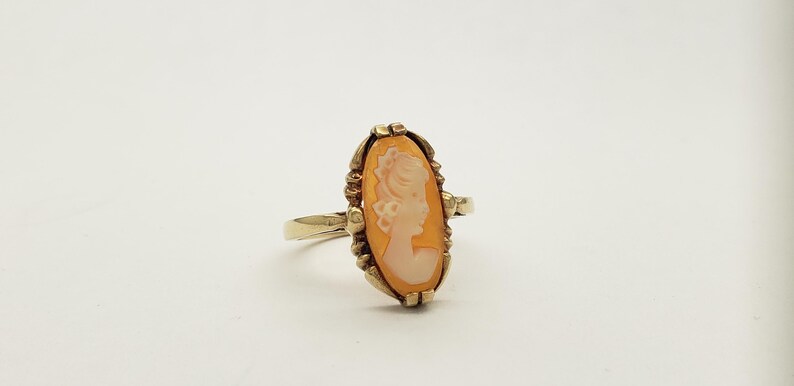 May include: Gold ring with a cameo featuring a woman's profile in a white cameo set in a gold oval frame.