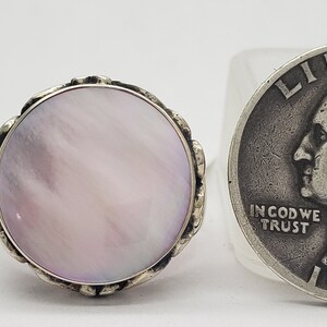 May include: A silver ring with a large, round, pink mother-of-pearl stone set in a decorative silver bezel. The ring is shown next to a US quarter for size comparison.