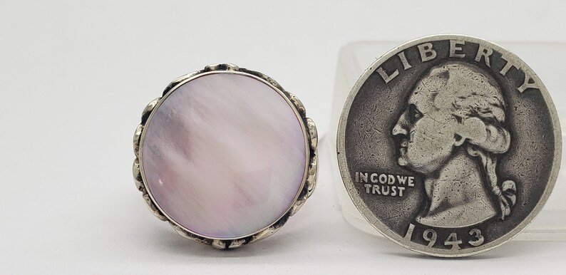 May include: A silver ring with a large, round, pink mother-of-pearl stone set in a decorative silver bezel. The ring is shown next to a US quarter for size comparison.