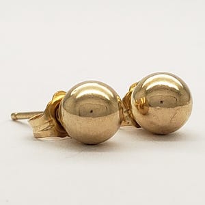Pu&ograve; includere: Un paio di orecchini a lobo a sfera color oro. Gli orecchini sono rotondi e lucenti, con perno e chiusura. Gli orecchini sono su uno sfondo bianco.