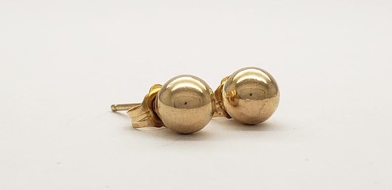 Pu&ograve; includere: Un paio di orecchini a lobo a sfera color oro. Gli orecchini sono rotondi e lucenti, con perno e chiusura. Gli orecchini sono su uno sfondo bianco.