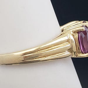 May include: A gold ring with a large, rectangular, purple gemstone. The ring has a ribbed design on the band and is set on a white surface against a dark background. The gemstone is a deep, rich color.