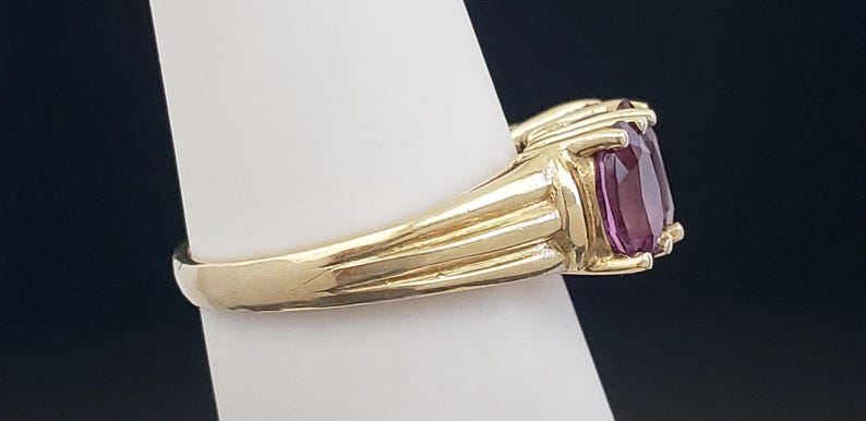 May include: A gold ring with a large, rectangular, purple gemstone. The ring has a ribbed design on the band and is set on a white surface against a dark background. The gemstone is a deep, rich color.