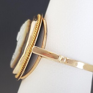May include: A gold ring featuring a cameo with a profile carving. The cameo is set in a dark oval frame, surrounded by a twisted gold rope design. The ring's band is a simple, polished gold.