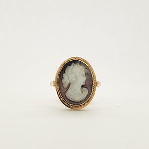 May include: Oval cameo ring featuring a profile of a woman with curly hair and a pearl necklace. The cameo is set in a gold-colored frame with a rope-like border. The ring has a simple band.