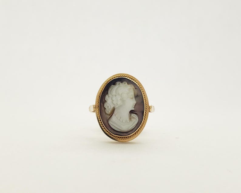 May include: Oval cameo ring featuring a profile of a woman with curly hair and a pearl necklace. The cameo is set in a gold-colored frame with a rope-like border. The ring has a simple band.