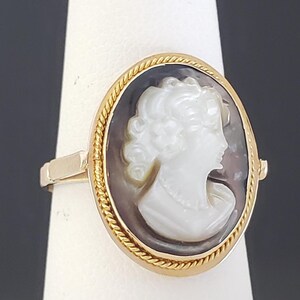 May include: Oval cameo ring featuring a white profile against a dark background, set in a gold-toned frame with a rope-like border. The ring's band is also gold-toned, showcasing a classic jewelry design.