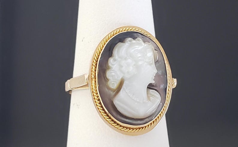 May include: Oval cameo ring featuring a white profile against a dark background, set in a gold-toned frame with a rope-like border. The ring's band is also gold-toned, showcasing a classic jewelry design.