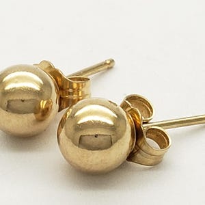 Pu&ograve; includere: Un paio di orecchini a lobo a sfera dorati. Ogni orecchino presenta un design sferico lucido. Gli orecchini hanno un perno e una chiusura a farfalla. Gli orecchini sono esposti su una superficie bianca.