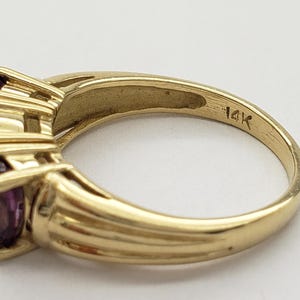 May include: A gold ring with three oval-cut purple gemstones. The ring is marked "14K" and has a ribbed design on the band. The gemstones are set in a prong setting.