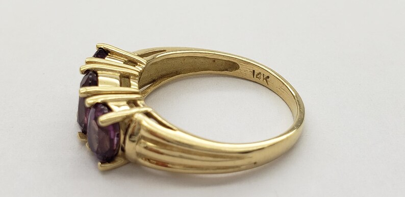 May include: A gold ring with three oval-cut purple gemstones. The ring is marked "14K" and has a ribbed design on the band. The gemstones are set in a prong setting.