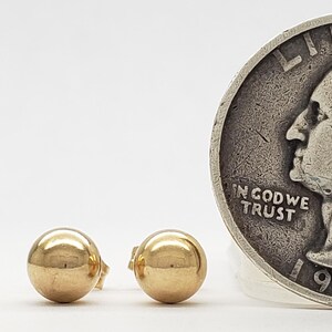 Pu&ograve; includere: Un paio di piccoli orecchini a bottone dorati a forma di sfera. Una moneta d'argento del 1935 &egrave; posizionata accanto agli orecchini per il confronto delle dimensioni. Gli orecchini sono rotondi e lucenti.