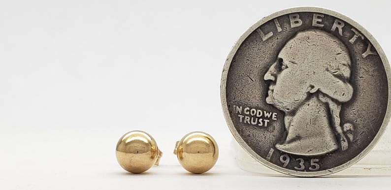 Pu&ograve; includere: Un paio di piccoli orecchini a bottone dorati a forma di sfera. Una moneta d'argento del 1935 &egrave; posizionata accanto agli orecchini per il confronto delle dimensioni. Gli orecchini sono rotondi e lucenti.