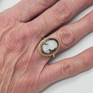 May include: A gold-toned ring featuring an oval cameo with a white profile against a dark background. The ring is set on a hand with visible skin texture, suggesting an antique or vintage piece. The ring's design is classic and elegant.