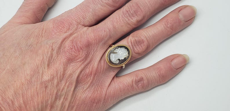 May include: A gold-toned ring featuring an oval cameo with a white profile against a dark background. The ring is set on a hand with visible skin texture, suggesting an antique or vintage piece. The ring's design is classic and elegant.