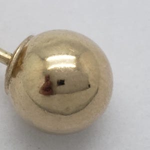 Pu&ograve; includere: Primo piano di un orecchino a sfera d'oro. L'orecchino presenta una piccola sfera rotonda d'oro lucido attaccata a un perno con chiusura a farfalla. L'oro riflette la luce, creando riflessi e ombre. L'orecchino &egrave; su sfondo bianco.