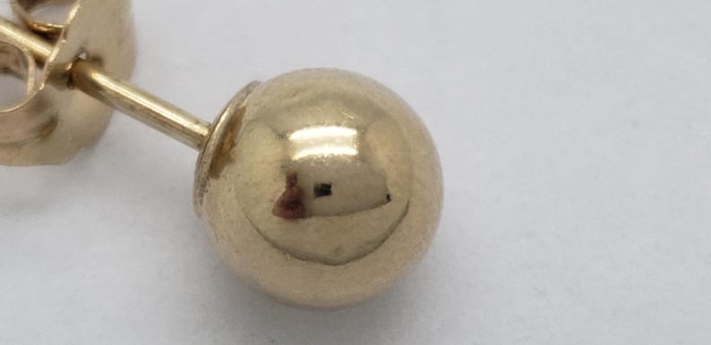 Pu&ograve; includere: Primo piano di un orecchino a sfera d'oro. L'orecchino presenta una piccola sfera rotonda d'oro lucido attaccata a un perno con chiusura a farfalla. L'oro riflette la luce, creando riflessi e ombre. L'orecchino &egrave; su sfondo bianco.