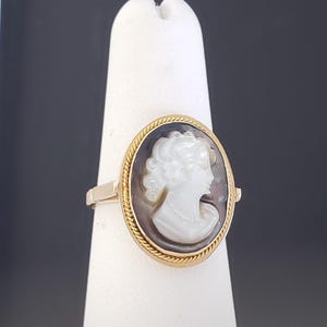 May include: Gold ring featuring an oval cameo with a white profile against a dark background. The cameo is framed by a gold rope-like border. The ring is displayed on a white stand against a dark background.