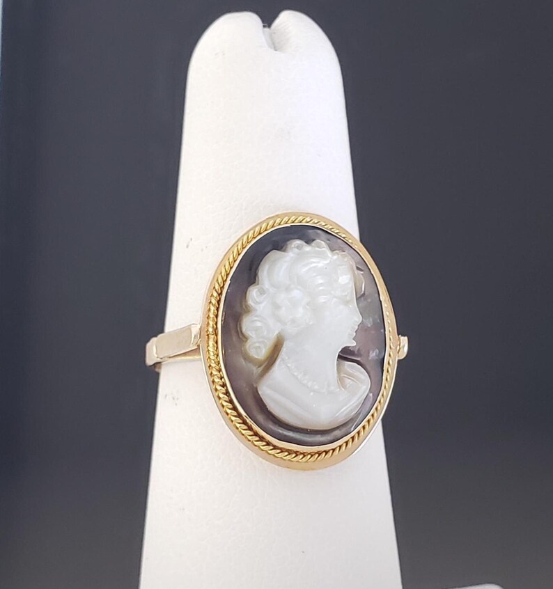 May include: Gold ring featuring an oval cameo with a white profile against a dark background. The cameo is framed by a gold rope-like border. The ring is displayed on a white stand against a dark background.