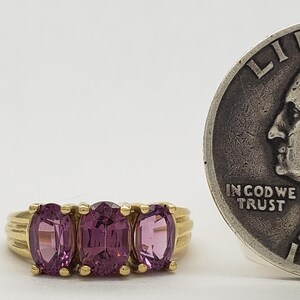 May include: A gold ring with three oval-cut, pink gemstones. A silver quarter coin is next to the ring. The coin has the inscription "IN GOD WE TRUST" and the year 1943.