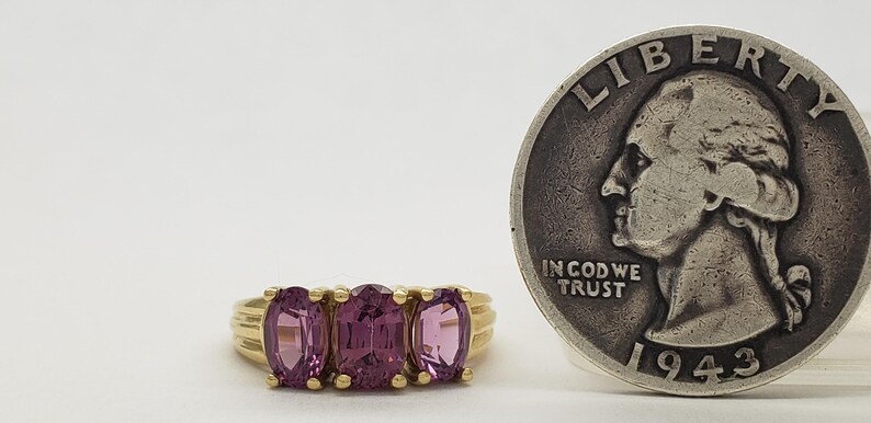 May include: A gold ring with three oval-cut, pink gemstones. A silver quarter coin is next to the ring. The coin has the inscription "IN GOD WE TRUST" and the year 1943.