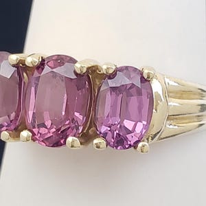 May include: A gold ring featuring three oval-cut, pink gemstones. The gemstones are set in a row, with a gold band and setting. The ring is displayed on a white surface against a dark background.