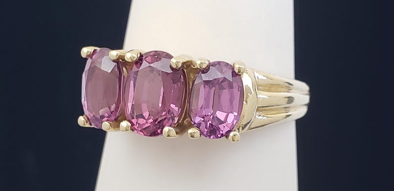 May include: A gold ring featuring three oval-cut, pink gemstones. The gemstones are set in a row, with a gold band and setting. The ring is displayed on a white surface against a dark background.