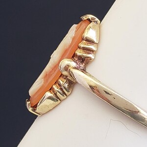 May include: A gold ring with a carved cameo set in the center. The cameo is a light orange color and has a detailed design.