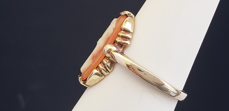 May include: A gold ring with a carved cameo set in the center. The cameo is a light orange color and has a detailed design.
