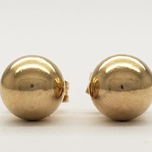 Pu&ograve; includere: Un paio di orecchini a lobo a sfera color oro. Gli orecchini sono rotondi e lucenti, riflettendo la luce. I perni sono visibili sul retro degli orecchini. Sono su sfondo bianco.