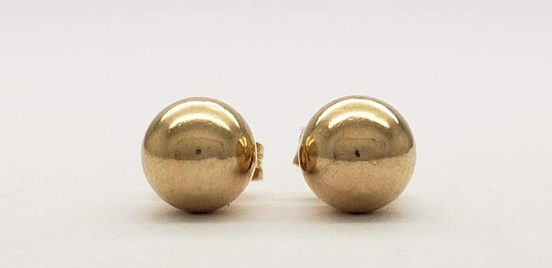 Pu&ograve; includere: Un paio di orecchini a lobo a sfera color oro. Gli orecchini sono rotondi e lucenti, riflettendo la luce. I perni sono visibili sul retro degli orecchini. Sono su sfondo bianco.