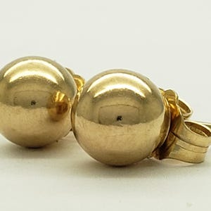 Pu&ograve; includere: Un paio di orecchini a bottone dorati. Gli orecchini hanno una superficie lucida e riflettente e un perno con chiusura a farfalla. Gli orecchini sono su uno sfondo bianco.