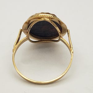 May include: A gold ring with a dark, oval-shaped stone. The ring has a detailed setting around the stone and a simple, rounded band. The ring is shown against a white background.