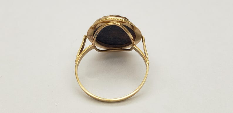 May include: A gold ring with a dark, oval-shaped stone. The ring has a detailed setting around the stone and a simple, rounded band. The ring is shown against a white background.