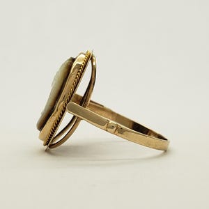 May include: A gold-toned ring featuring a cameo design. The ring has a detailed oval cameo set within a decorative frame. The band and frame have a polished finish, creating a classic and elegant look. The ring is shown from a side angle.