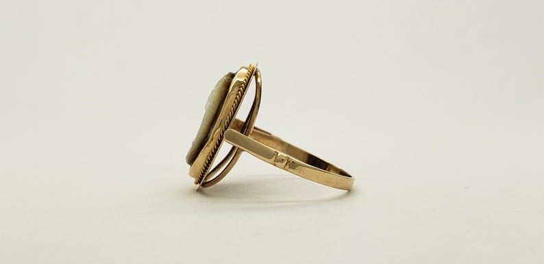 May include: A gold-toned ring featuring a cameo design. The ring has a detailed oval cameo set within a decorative frame. The band and frame have a polished finish, creating a classic and elegant look. The ring is shown from a side angle.