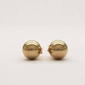 Pu&ograve; includere: Due orecchini a bottone sferici color oro. Ogni orecchino ha una superficie liscia e riflettente e una chiusura a perno e chiusura. Gli orecchini sono esposti su sfondo bianco.