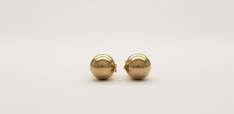 Pu&ograve; includere: Due orecchini a bottone sferici color oro. Ogni orecchino ha una superficie liscia e riflettente e una chiusura a perno e chiusura. Gli orecchini sono esposti su sfondo bianco.