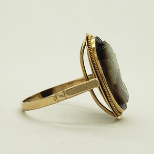 May include: A gold ring featuring a cameo with a profile portrait. The cameo is set in an oval frame with a rope-like border. The ring's band and setting are a polished gold color.