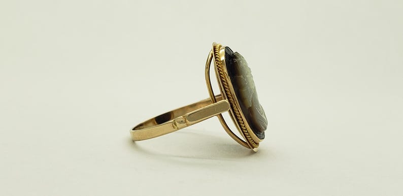 May include: A gold ring featuring a cameo with a profile portrait. The cameo is set in an oval frame with a rope-like border. The ring's band and setting are a polished gold color.