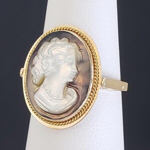 May include: A gold ring featuring a cameo with a profile of a woman. The oval cameo is set within a gold frame with a twisted rope design. The woman's profile is carved in white, set against a brown background.