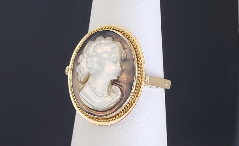 May include: A gold ring featuring a cameo with a profile of a woman. The oval cameo is set within a gold frame with a twisted rope design. The woman's profile is carved in white, set against a brown background.