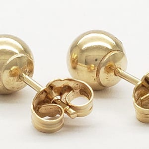 Pu&ograve; includere: Un paio di orecchini a lobo dorati a forma di sfera. Ogni orecchino presenta un design sferico liscio con un perno e una chiusura a farfalla. Gli orecchini sono esposti su una superficie bianca.