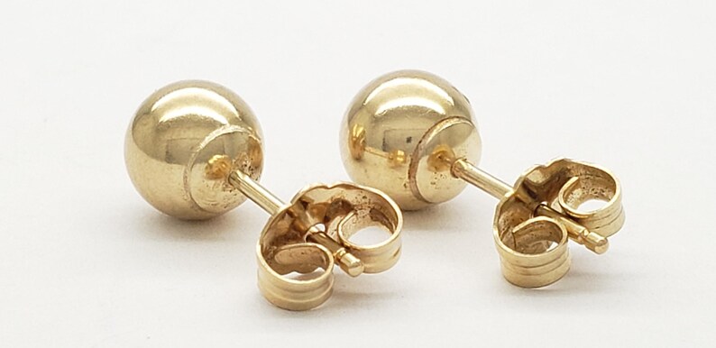 Pu&ograve; includere: Un paio di orecchini a lobo dorati a forma di sfera. Ogni orecchino presenta un design sferico liscio con un perno e una chiusura a farfalla. Gli orecchini sono esposti su una superficie bianca.