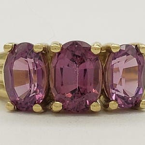 May include: A gold ring featuring three oval-cut, pink gemstones. The gemstones are set in a row, with a gold band that has a ribbed design on the sides. The ring is displayed against a white background.