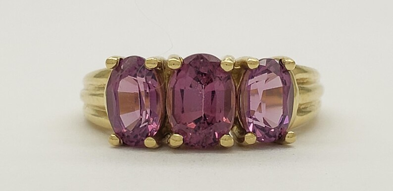 May include: A gold ring featuring three oval-cut, pink gemstones. The gemstones are set in a row, with a gold band that has a ribbed design on the sides. The ring is displayed against a white background.