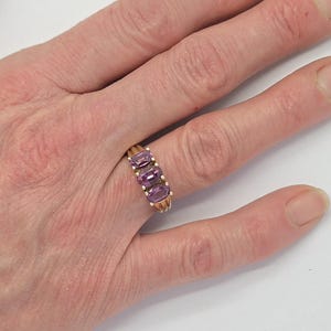 May include: A gold-toned ring with three oval, purple gemstones set in a row. The ring is on a person's hand, with the fingers slightly spread. The background is plain white.