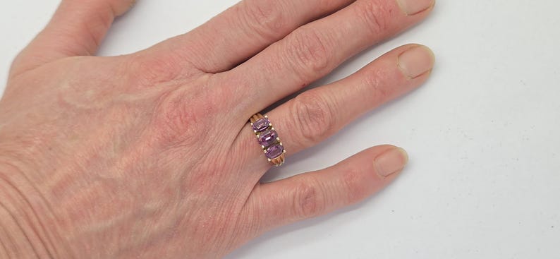 May include: A gold-toned ring with three oval, purple gemstones set in a row. The ring is on a person's hand, with the fingers slightly spread. The background is plain white.