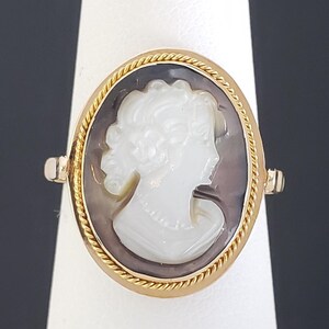 May include: Gold ring featuring an oval cameo with a white profile against a dark background. The cameo is framed by a twisted gold rope design. The ring is set on a white stand against a dark background.